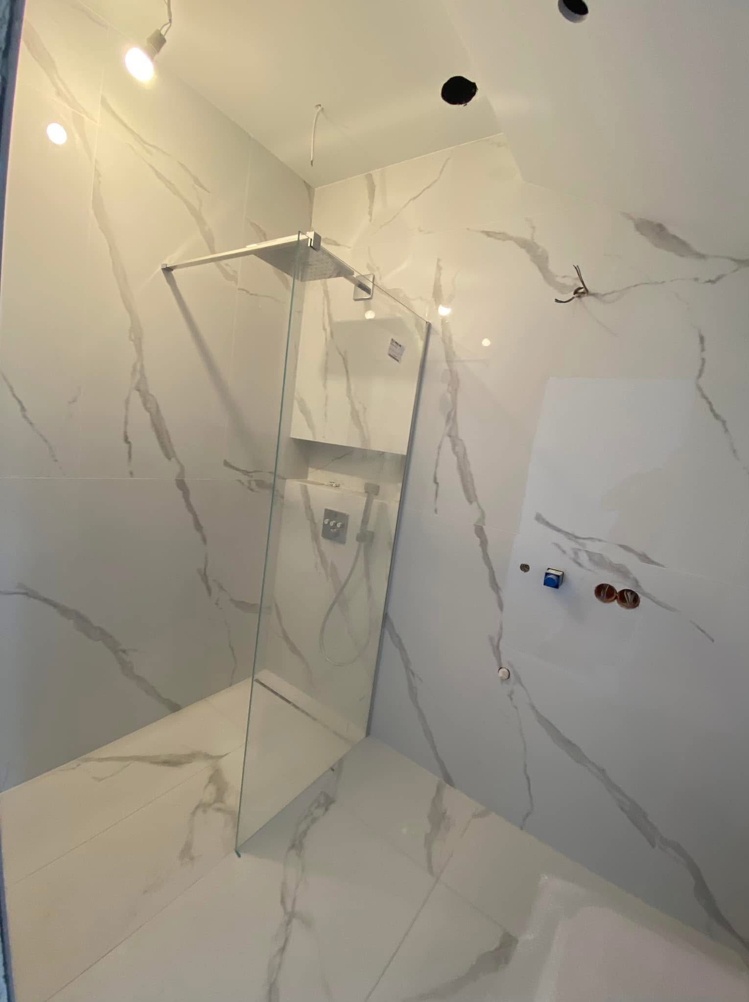 Nowoczesna kabina prysznicowa walk-in z białymi płytkami imitującymi marmur, widoczne niedokończone prace elektryczne na ścianie, surowe oświetlenie punktowe.