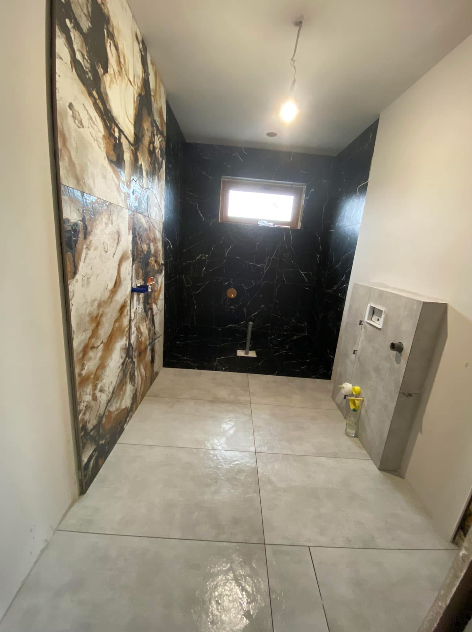 Wnętrze łazienki w trakcie wykańczania, z widocznymi płytkami imitującymi marmur na ścianie, szarą posadzką i fragmentem zabudowy z szarej płyty. Uwagę zwraca kontrast między jasnymi i ciemnymi...