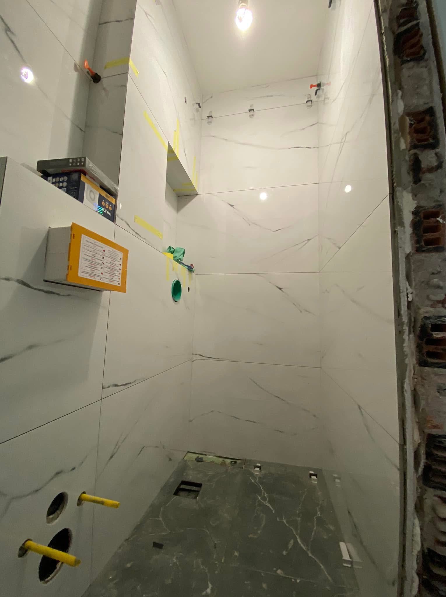 Wnętrze łazienki w trakcie prac wykończeniowych, widoczne białe płytki imitujące marmur na ścianach, szare płytki na podłodze, otwory na instalacje sanitarne z wystającymi żółtymi rurkami, fragment...