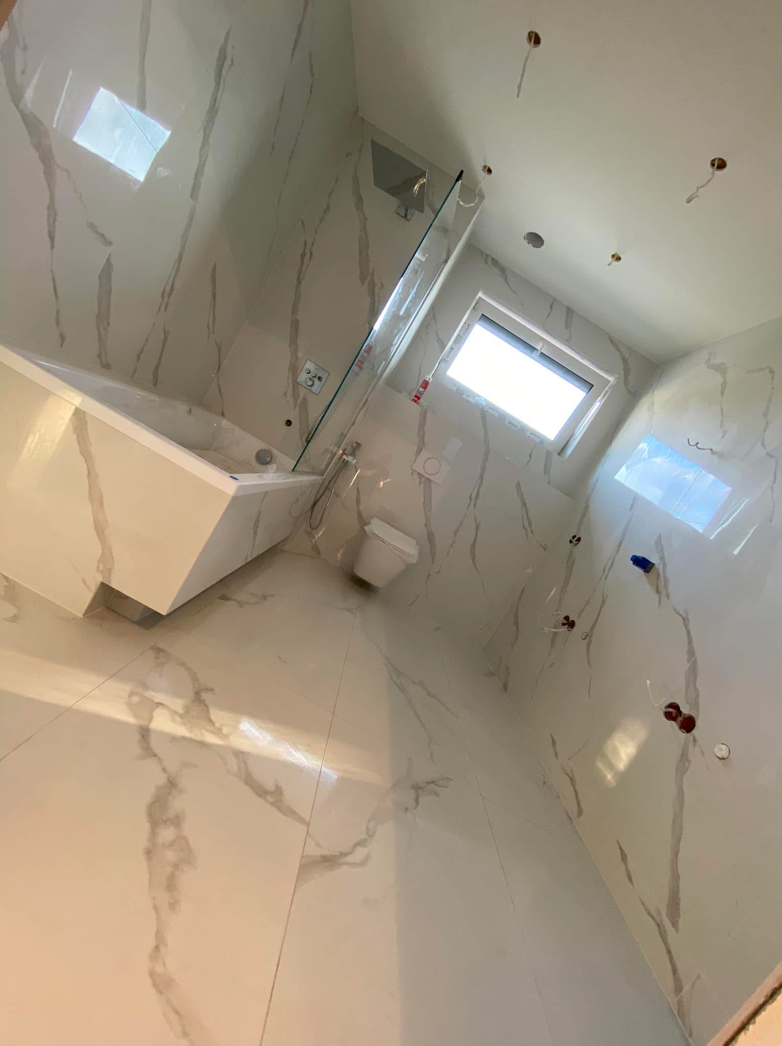 Nowoczesna, jasna łazienka z wanną obudowaną marmurem, szklanym panelem prysznicowym i toaletą. Ściany i podłoga wyłożone płytkami imitującymi biały marmur z szarymi żyłami. Widoczne przygotowane...