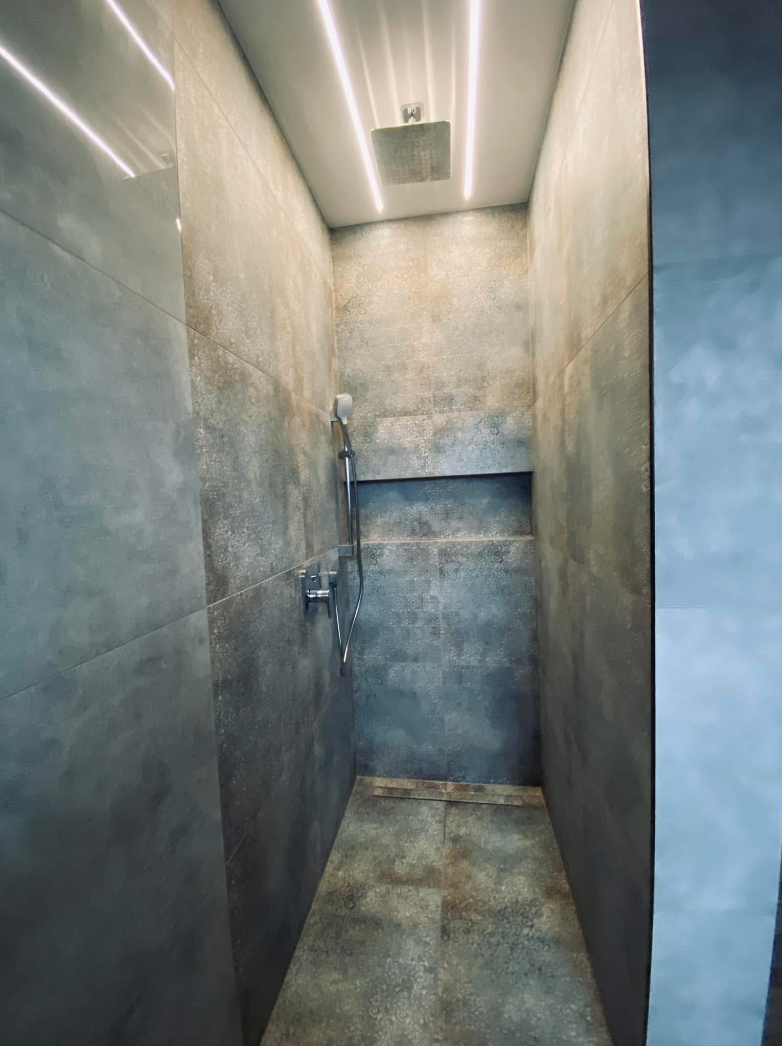 Wnętrze kabiny prysznicowej wykończonej szarymi płytkami imitującymi beton, z wnęką na kosmetyki i oświetleniem LED w suficie.