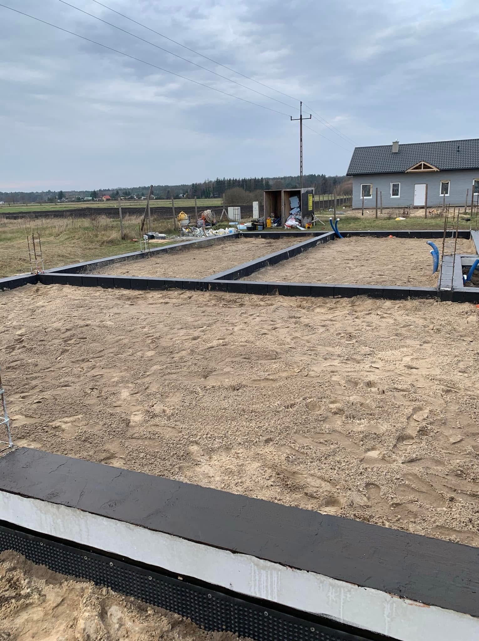 Przygotowany fundament pod budynek jednorodzinny: czarna hydroizolacja pionowa na styropianie, piasek zasypowy, zbrojenie pionowe, niebieskie rury instalacyjne, w tle dom w szarej elewacji...