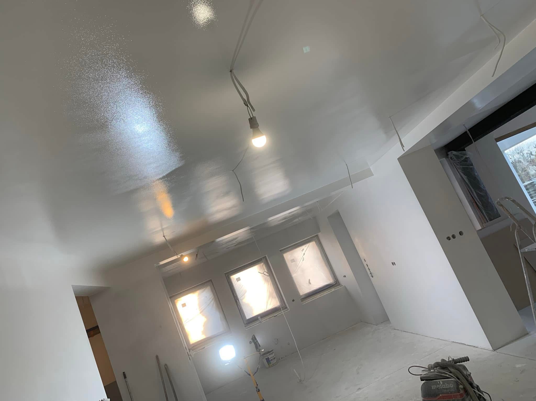 Świeżo położona, błyszcząca gładź na suficie w trakcie schnięcia, widoczne tymczasowe oświetlenie, zakryte okna i sprzęt do szlifowania na podłodze.