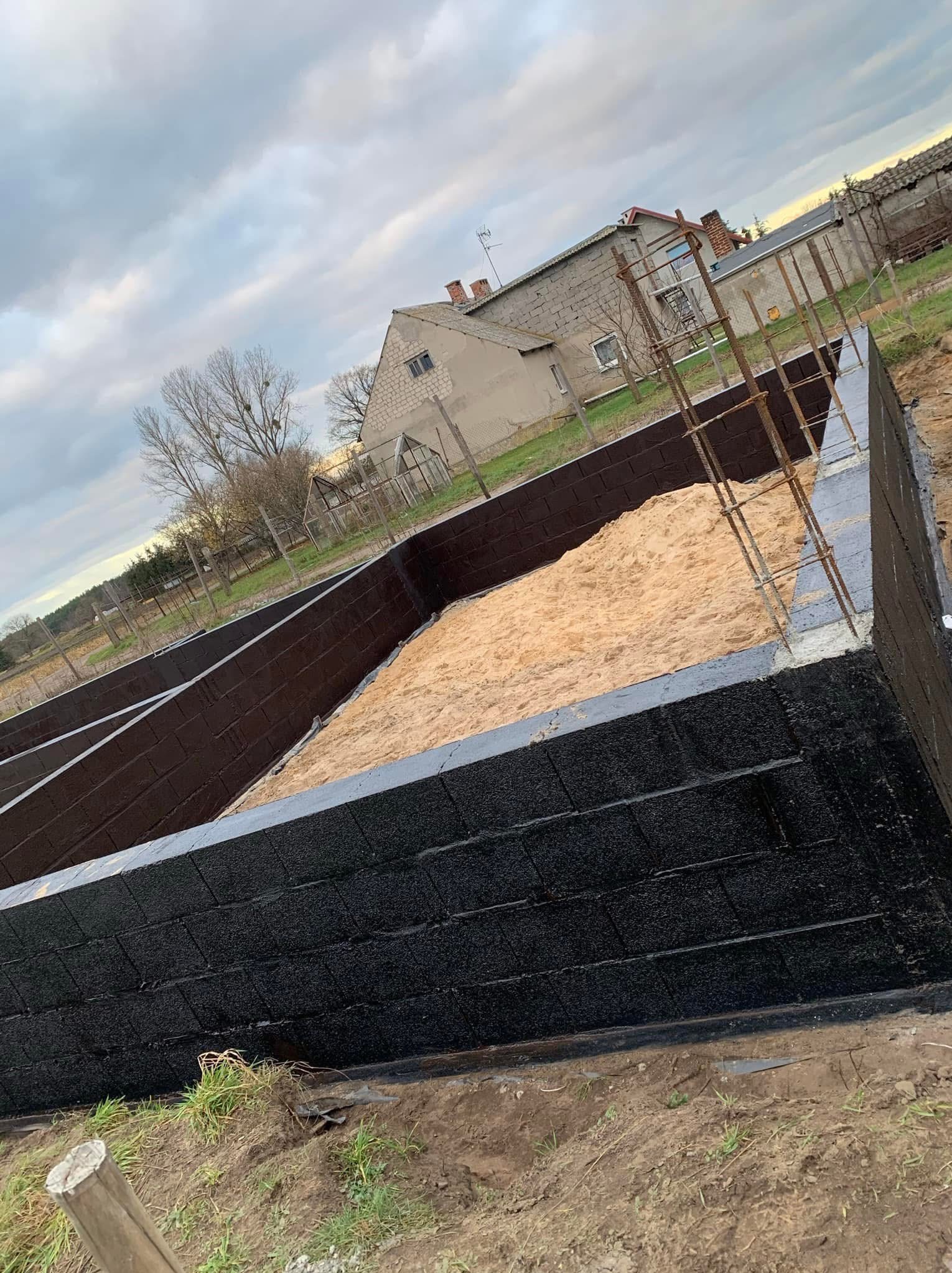 Fundamenty budynku z bloczków betonowych, zabezpieczone czarną izolacją, częściowo zasypane piaskiem, widoczne zbrojenia, w tle budynki gospodarcze i drzewa.