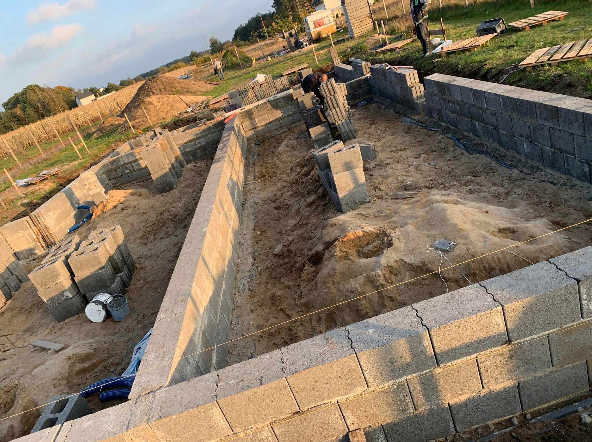 Budowa fundamentów domu jednorodzinnego z bloczków betonowych, widoczny wykop, ułożone pierwsze warstwy bloczków, poziomowanie sznurkiem, zapas bloczków na paletach i wiaderka z zaprawą wewnątrz...