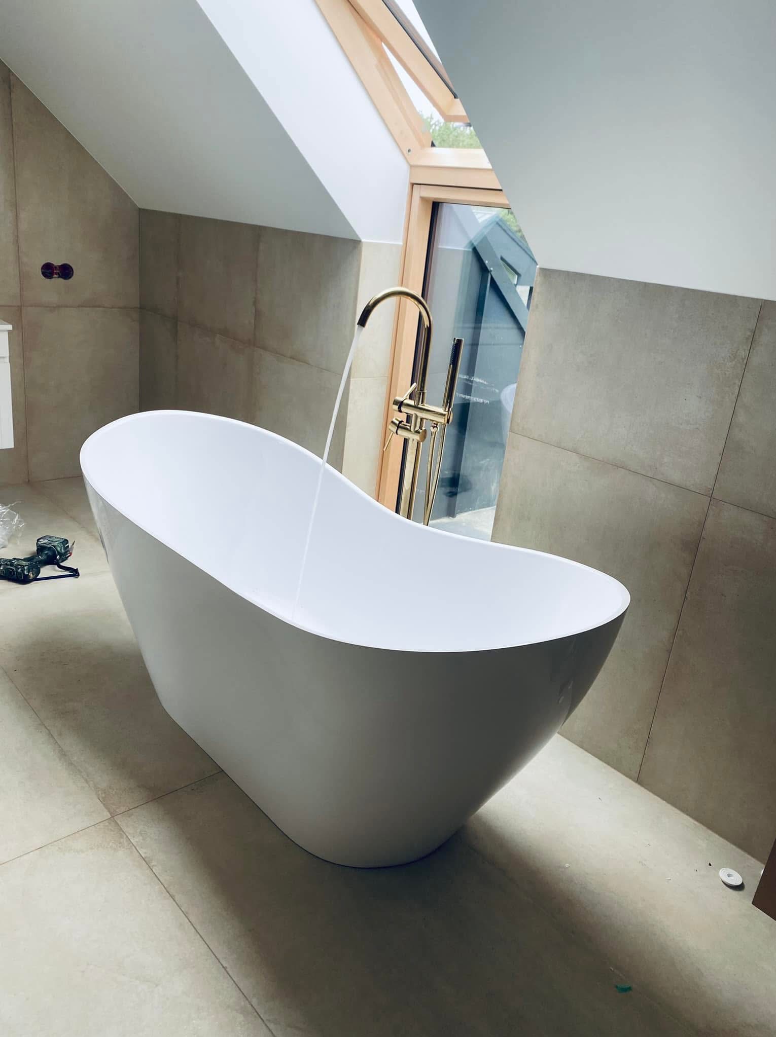 Biała, wolnostojąca wanna z złotą armaturą na tle beżowych płytek i okna dachowego, widoczne narzędzia na podłodze wskazujące na trwający montaż.