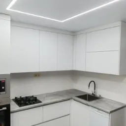 Nowoczesna, minimalistyczna kuchnia w bieli z szarym blatem w Warszawie. Widoczny piekarnik, płyta gazowa, szafki wiszące i oświetlenie LED w suficie.