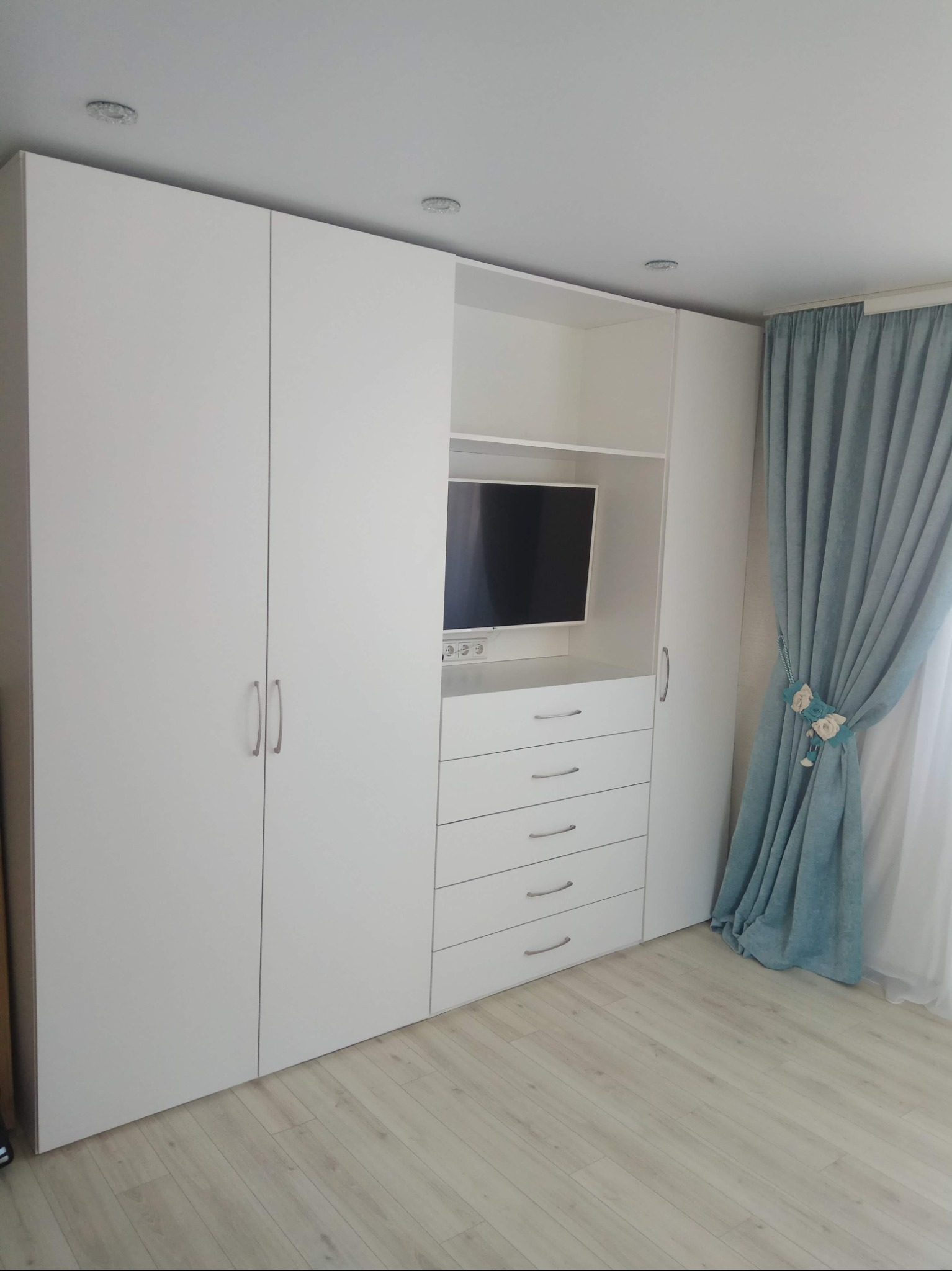 Biała szafa wnękowa z telewizorem wbudowanym w środkową część, obok okna z turkusowymi zasłonami z dekoracjami kwiatowymi.