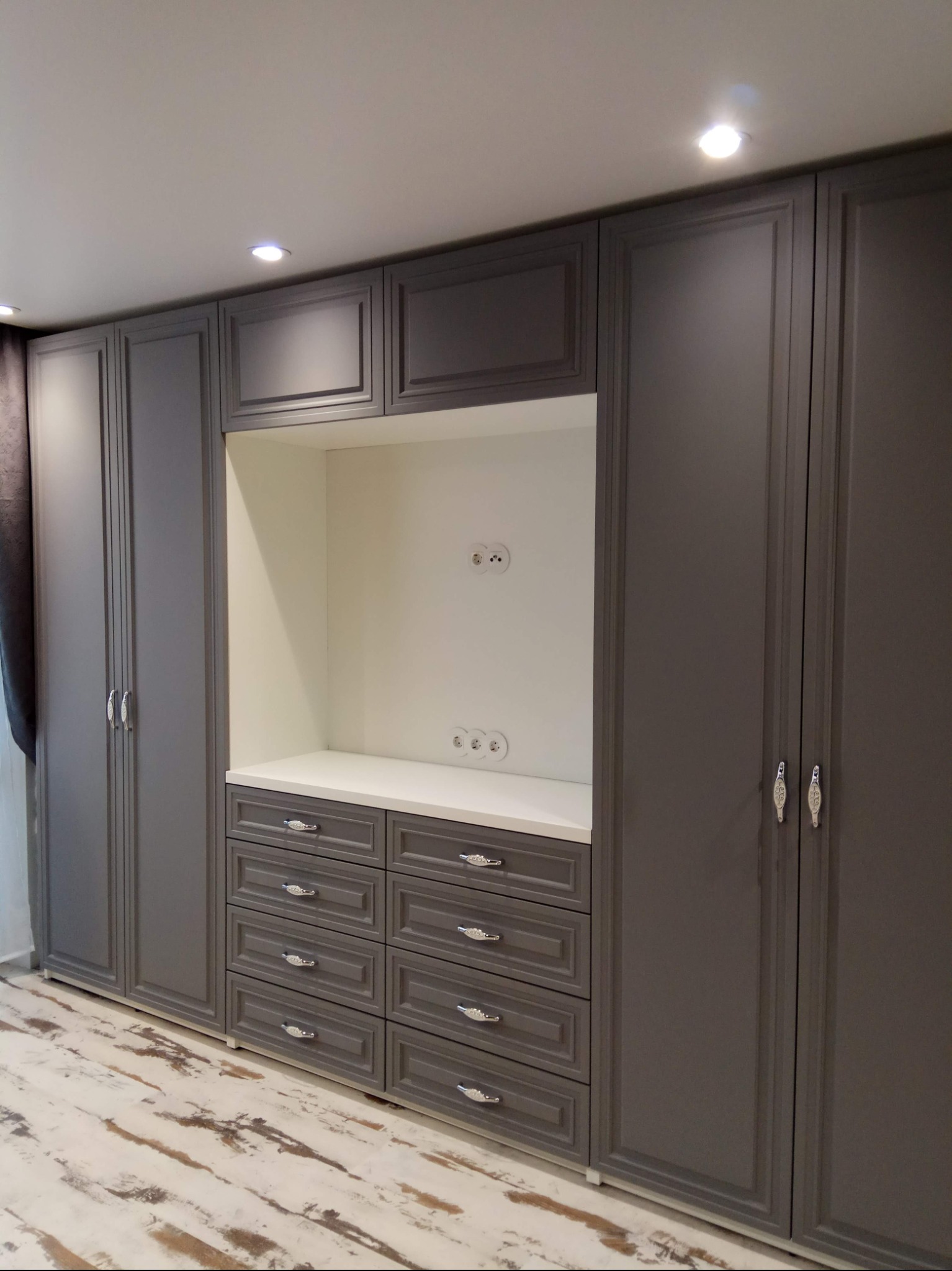 Szara zabudowa meblowa z szafami i szufladami, z wnęką na sprzęt RTV z białym wnętrzem i gniazdkami, podłoga imitująca drewno.