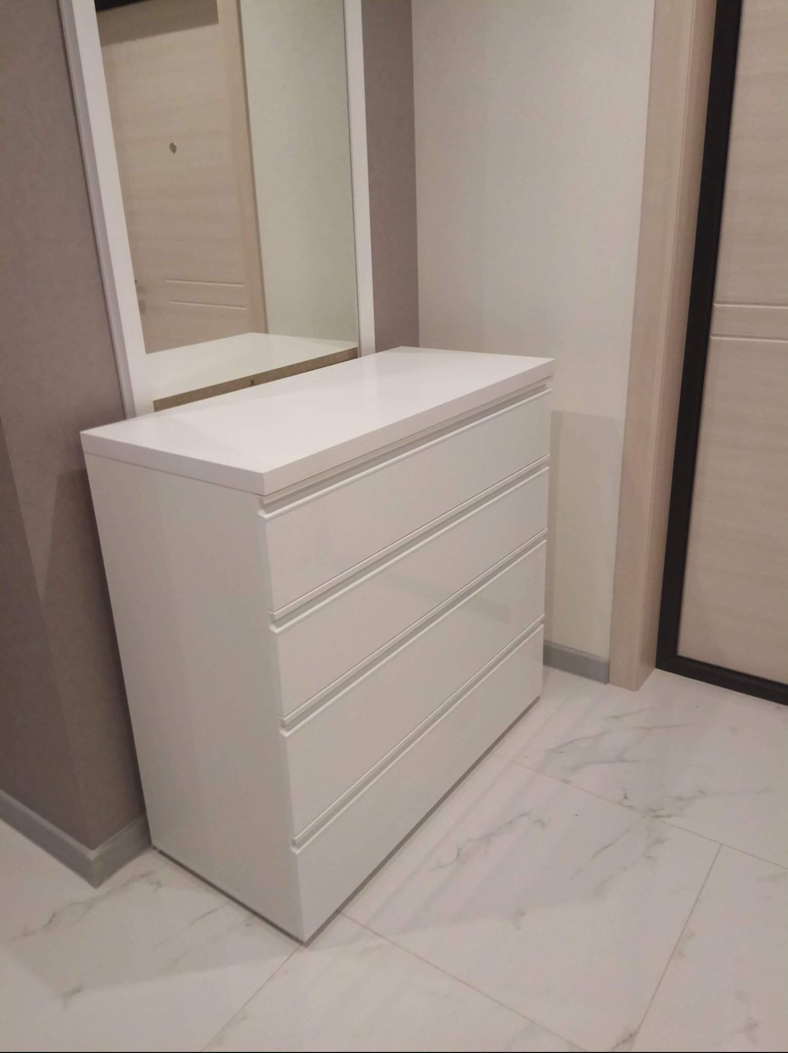 Biała komoda z trzema szufladami i lustrem w białej ramie, widok z boku, marmurowa podłoga.