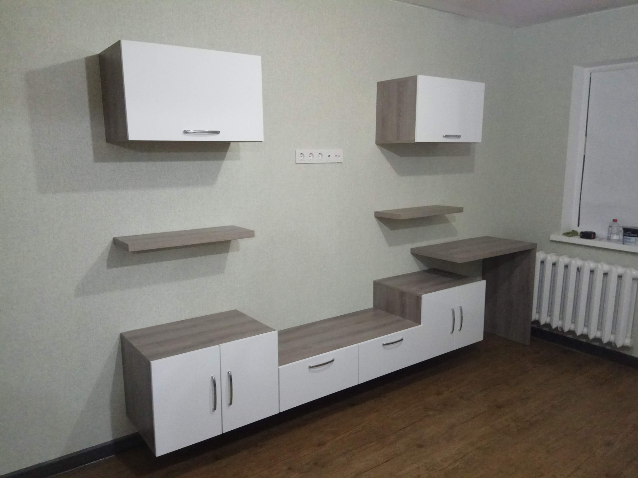 Nowoczesna zabudowa ściany z szafkami wiszącymi i dolnymi w kolorze białym i dębowym, z drewnianą podłogą i grzejnikiem żeliwnym pod oknem.