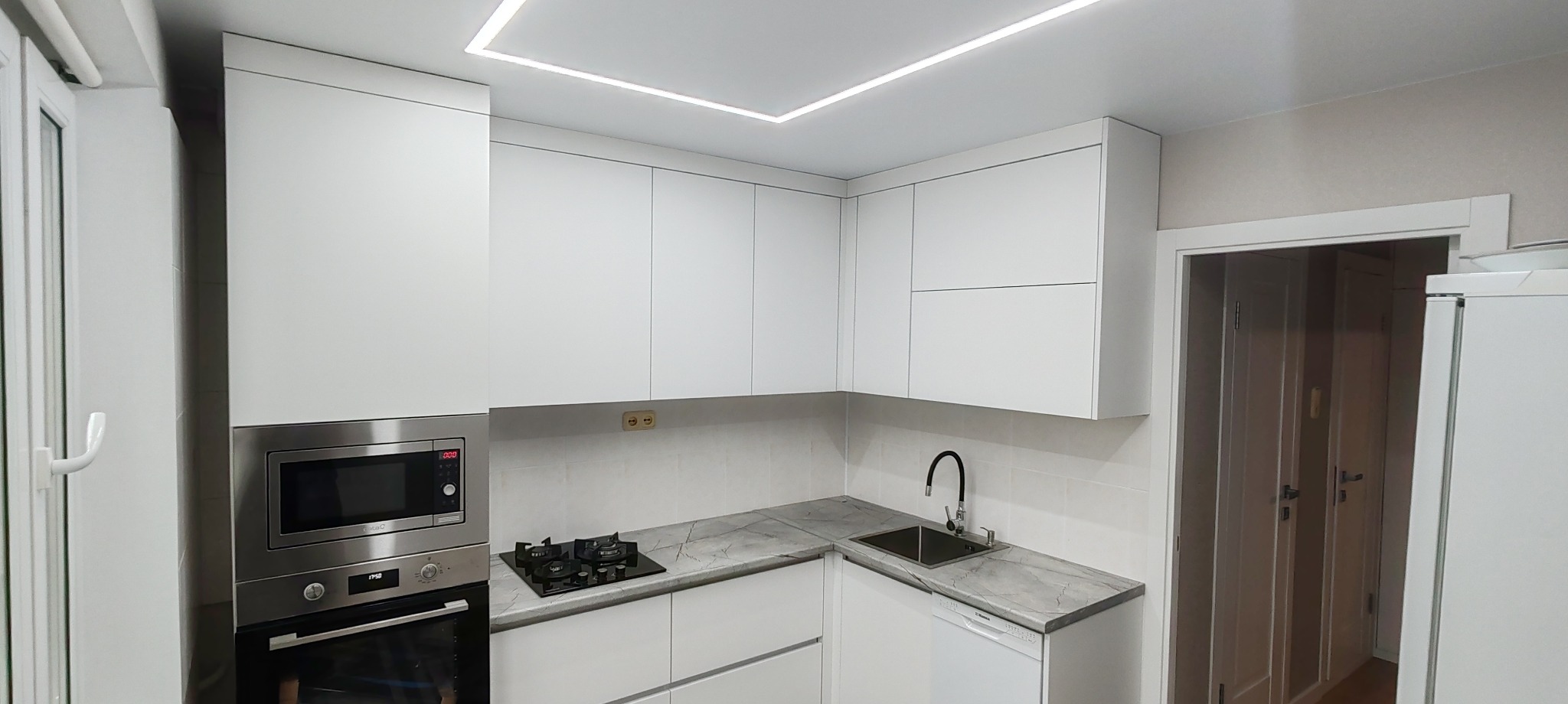 Nowoczesna, minimalistyczna kuchnia w bieli z szarym blatem w Warszawie. Widoczny piekarnik, płyta gazowa, szafki wiszące i oświetlenie LED w suficie.