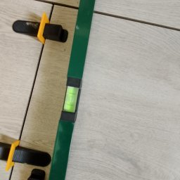 Czapkbud - Układanie płytek podłogowych imitujących drewno, z użyciem poziomicy i klipsów do poziomowania, widoczne fugi.