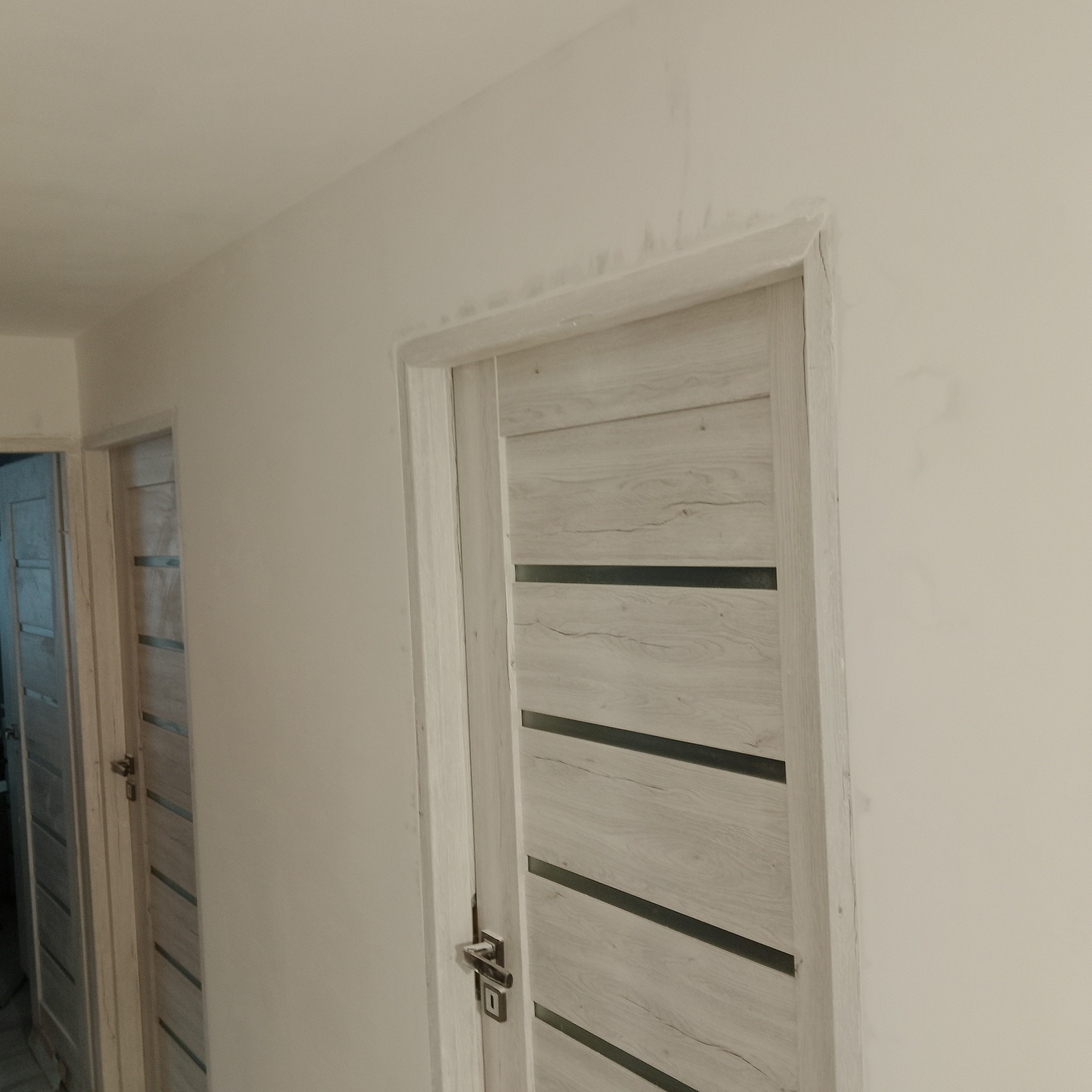 Zamontowane drzwi wewnętrzne w odcieniach bieli z poziomymi czarnymi paskami, widoczne na tle świeżo pomalowanej ściany i sufitu z delikatnymi niedociągnięciami w wykończeniu.