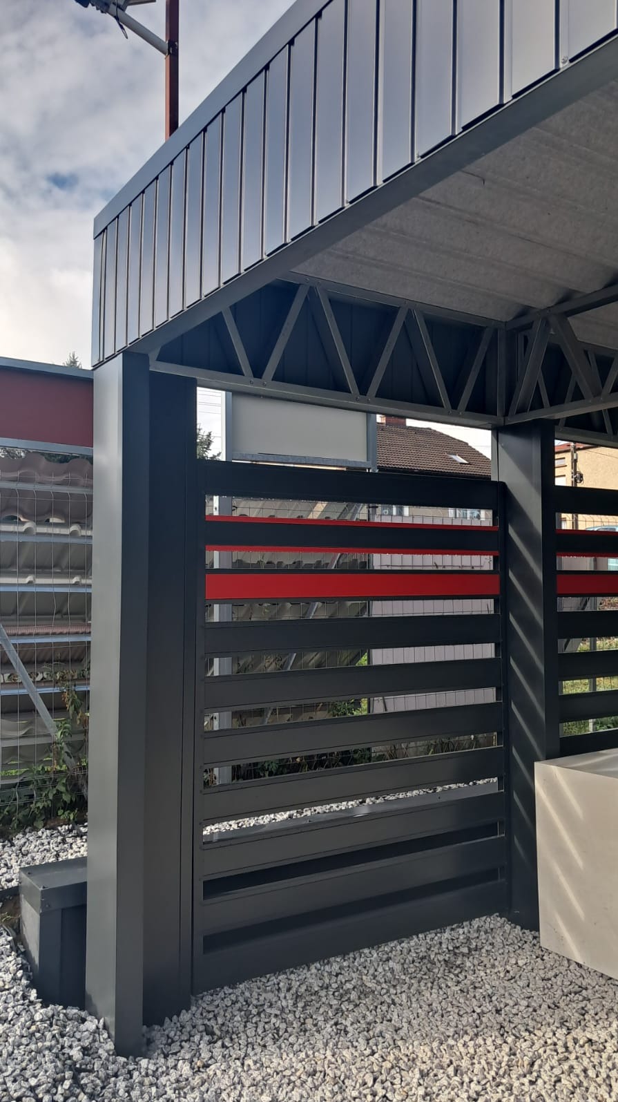 Nowoczesny carport z grafitową konstrukcją, poziomymi panelami z akcentem czerwieni i zadaszeniem z blachy profilowanej, otoczony białym żwirem.