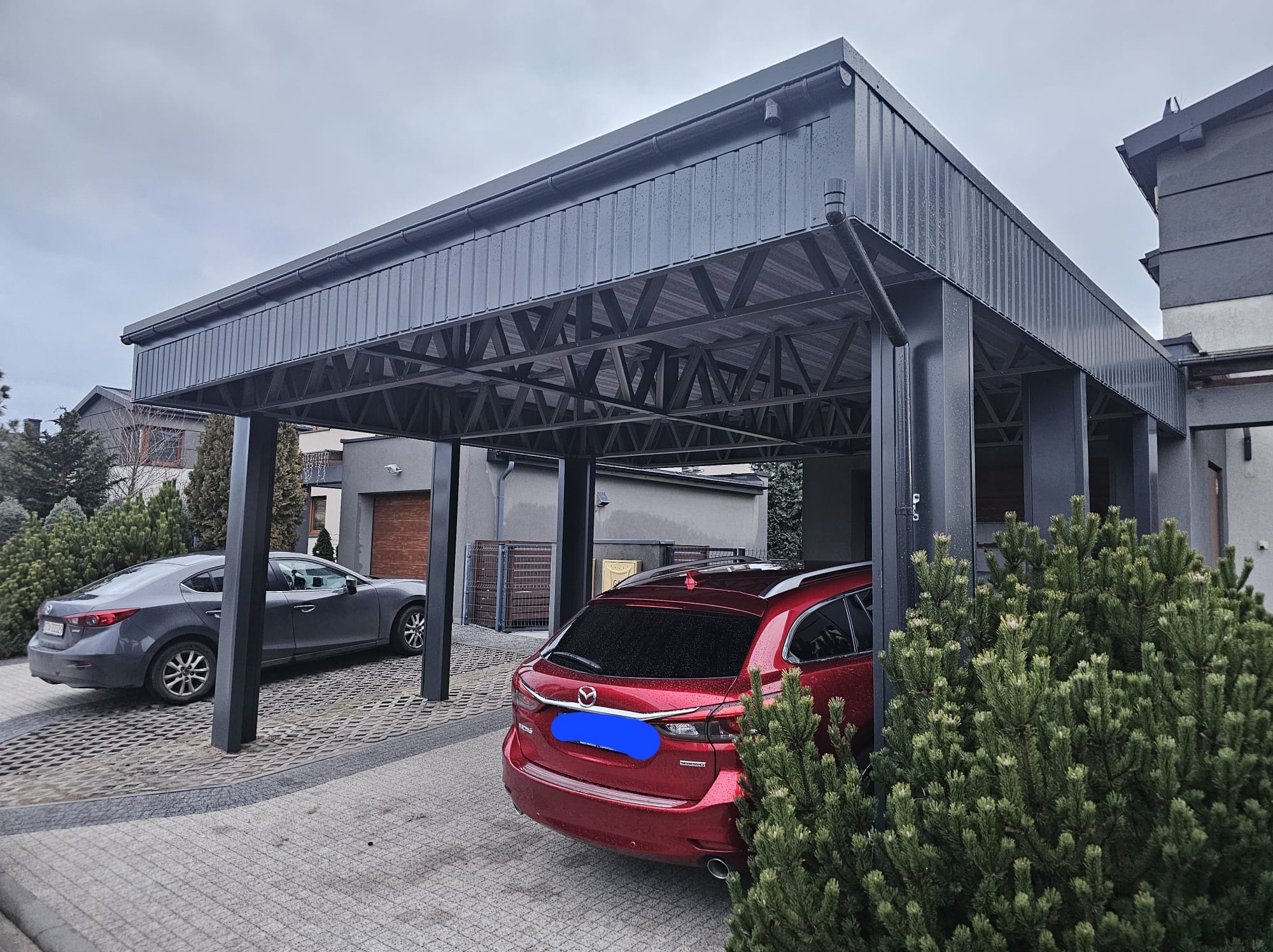 Nowoczesny, grafitowy carport z blachy trapezowej, z widoczną konstrukcją nośną, chroniący dwa zaparkowane samochody Mazda na posesji z kostki brukowej.