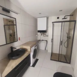 Nowoczesna łazienka w Żorach z kabiną prysznicową typu walk-in z czarnymi profilami, umywalką nablatową w kolorze czarnym, pralką oraz grzejnikiem, wykończona białymi płytkami.