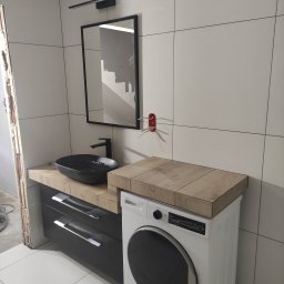 Decor-Marc Przemysław Marcinkowski - Nowoczesna łazienka w trakcie wykańczania: czarna umywalka nablatowa, szafka z szufladami, pralka z drewnopodobnym blatem, białe płytki ścienne, widoczne kable elektryczne i niedokończone obrzeże.