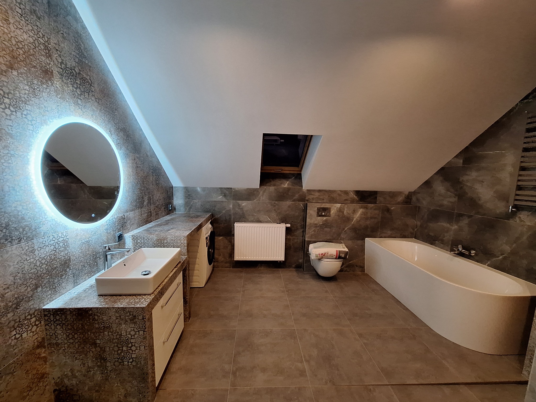 Nowoczesna łazienka na poddaszu z płytkami imitującymi marmur, okrągłym lustrem z podświetleniem LED, białą umywalką nablatową, wanną wolnostojącą i toaletą z papierem, wykończona w szarościach...