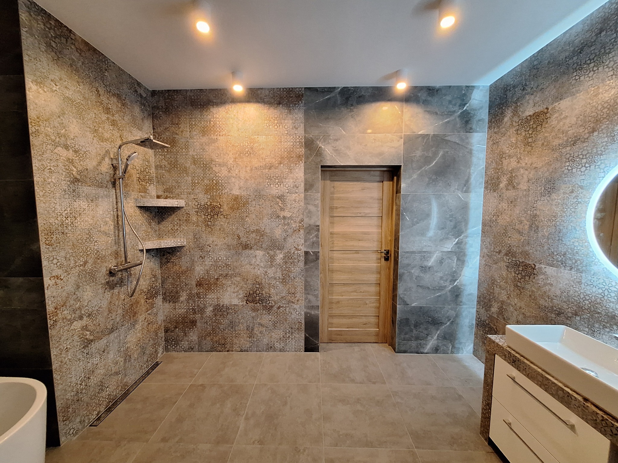 Nowoczesna łazienka z kabiną prysznicową typu walk-in, wykończona płytkami w odcieniach beżu i szarości, z drewnianymi drzwiami i podświetlanym lustrem nad umywalką.