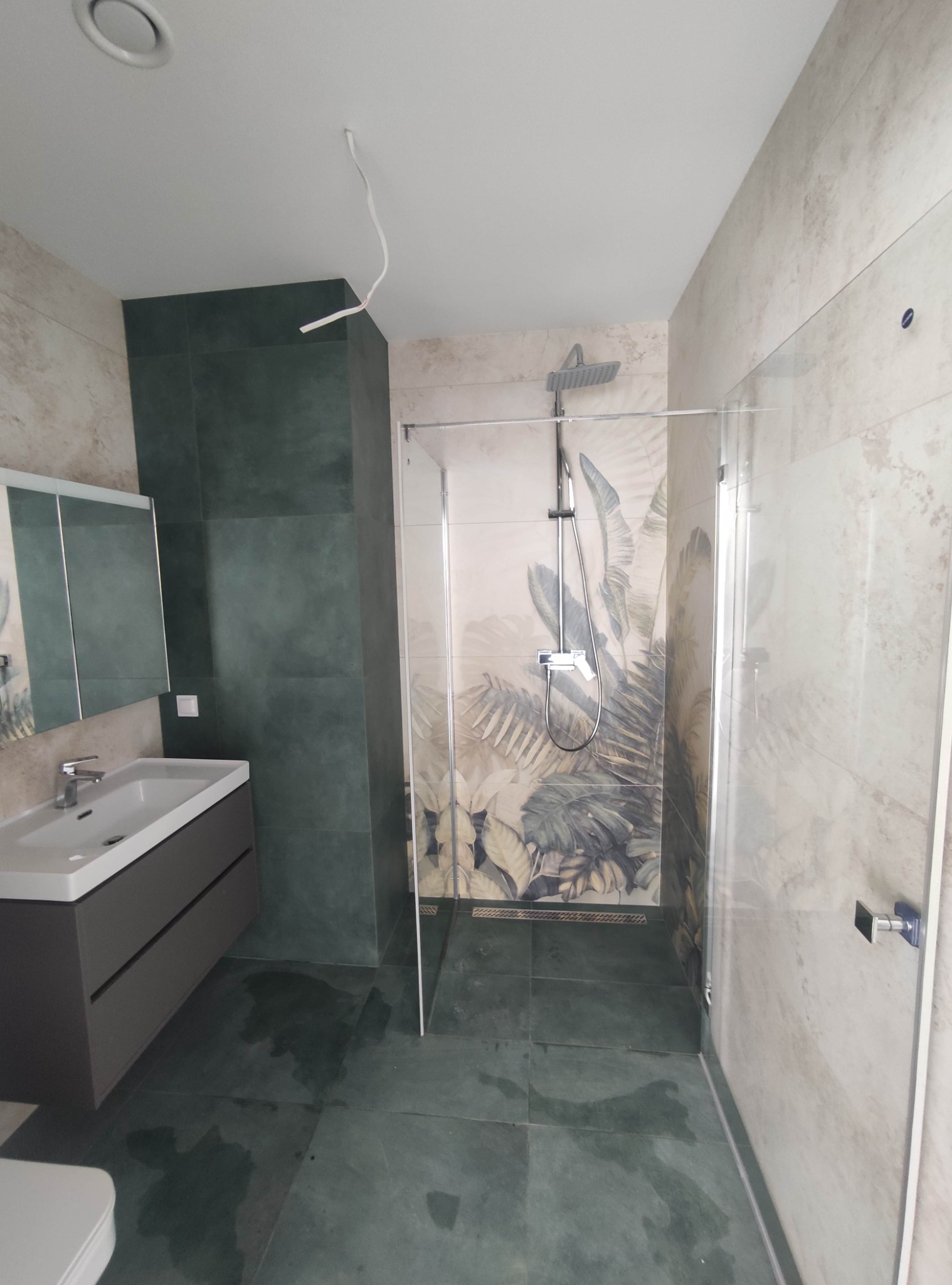 Nowoczesna łazienka z kabiną prysznicową typu walk-in, umywalką z szafką w kolorze grafitowym, oraz zielonymi płytkami imitującymi kamień na ścianach i podłodze; na ścianie pod prysznicem płytki...