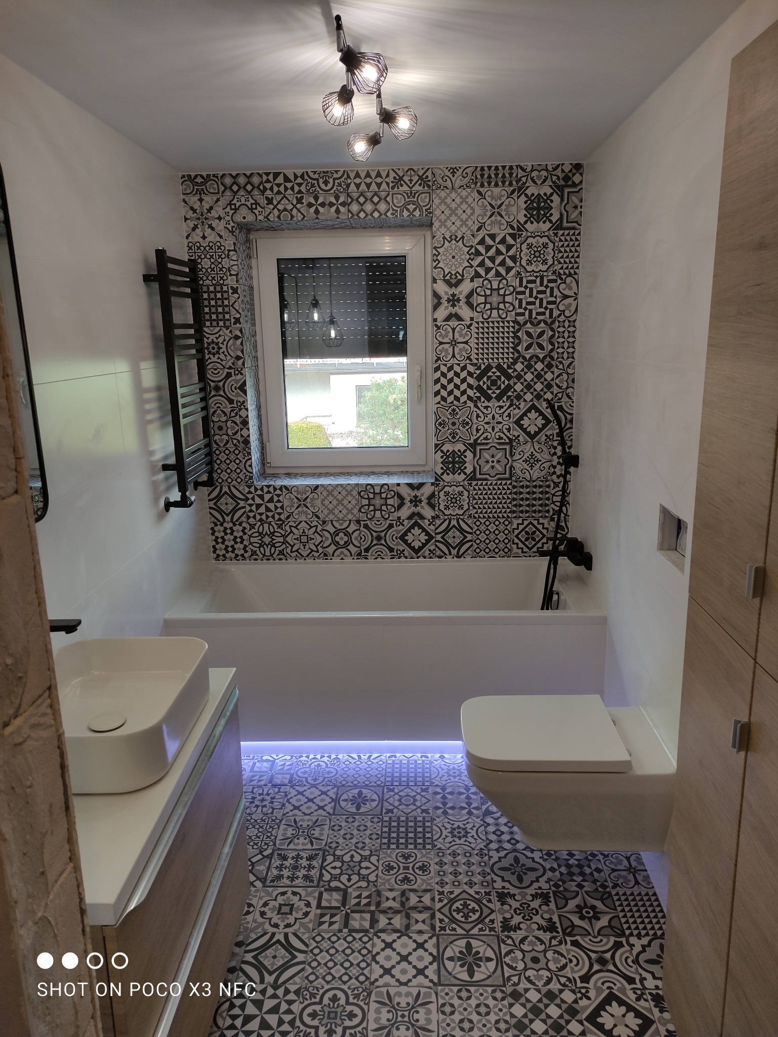 Wykończona łazienka z wzorzystymi, czarno-białymi płytkami podłogowymi i ściennymi, podświetleniem LED pod wanną, wiszącą toaletą i umywalką nablatową.