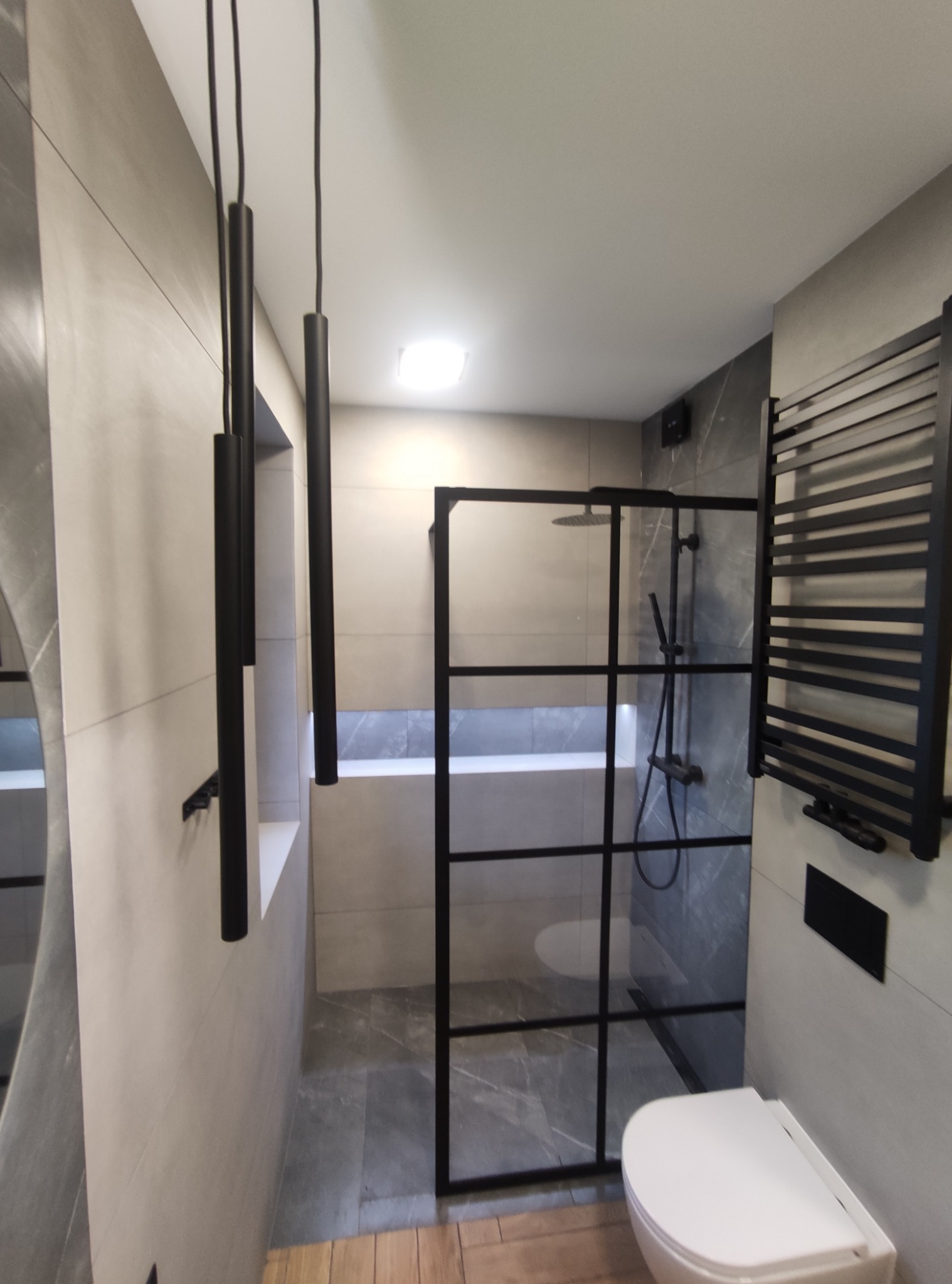 Nowoczesna łazienka z kabiną prysznicową typu walk-in z czarnymi profilami, wiszącą toaletą, grzejnikiem drabinkowym w czarnym kolorze i podwieszanymi lampami rurowymi