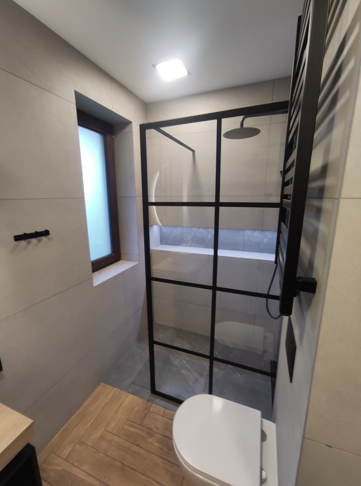 Nowoczesna łazienka z kabiną prysznicową w stylu loft z czarnymi profilami, grzejnikiem drabinkowym i płytkami imitującymi drewno na podłodze.