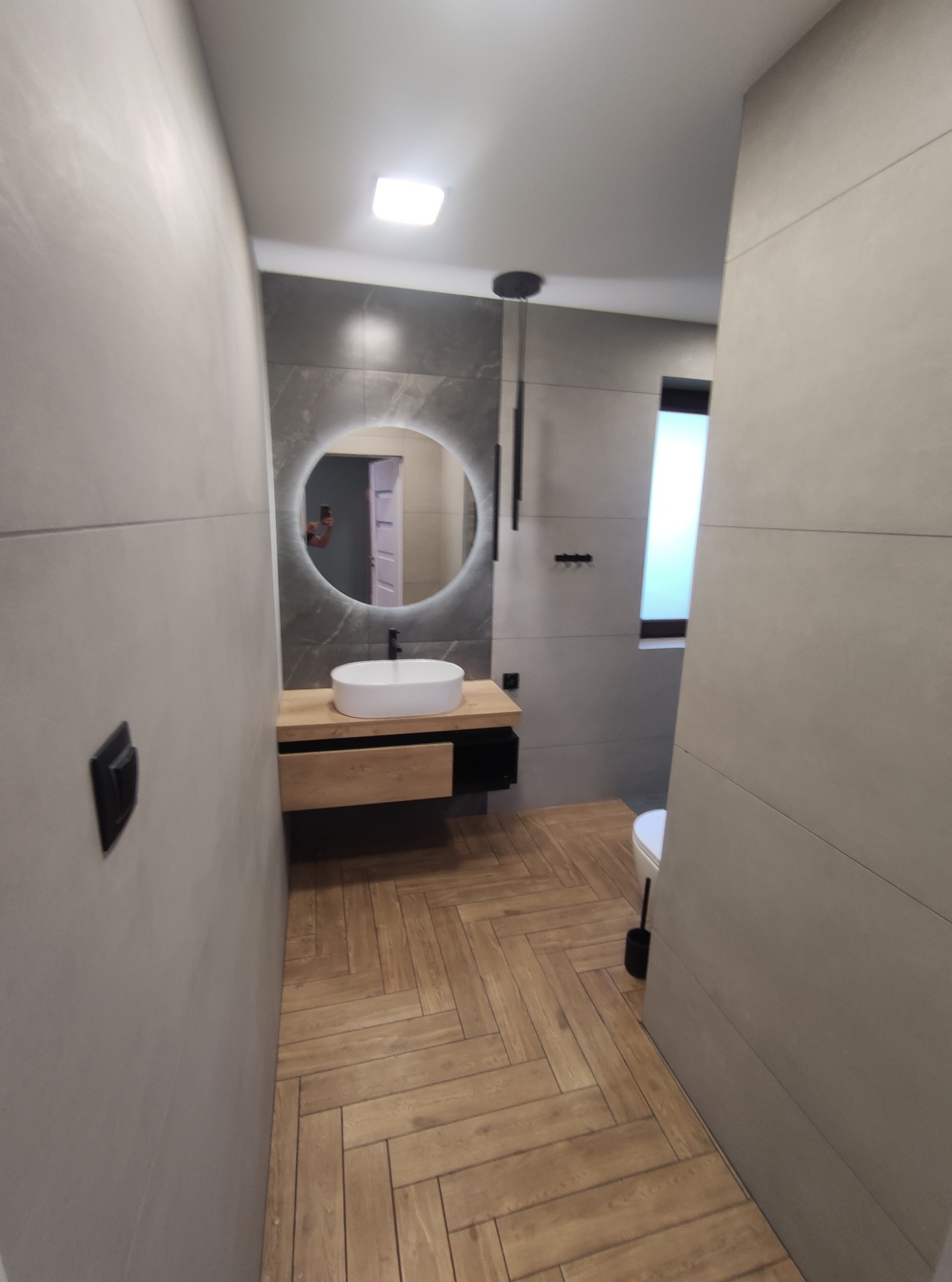 Nowoczesna łazienka z drewnianą podłogą ułożoną w jodełkę, okrągłym lustrem z podświetleniem LED, umywalką nablatową i szarą armaturą.