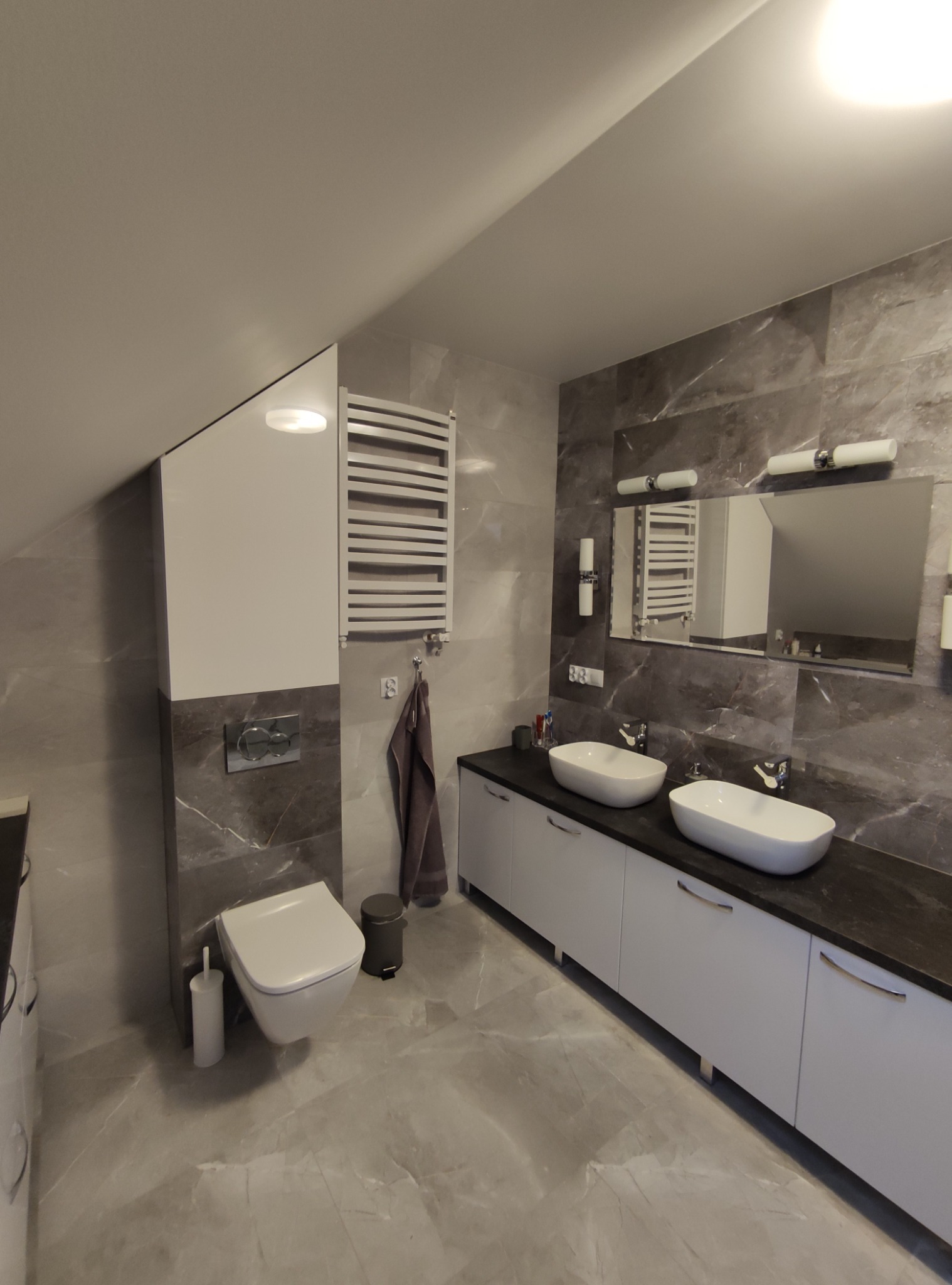 Nowoczesna łazienka na poddaszu z marmurowymi płytkami, umywalkami nablatowymi i grzejnikiem drabinkowym.