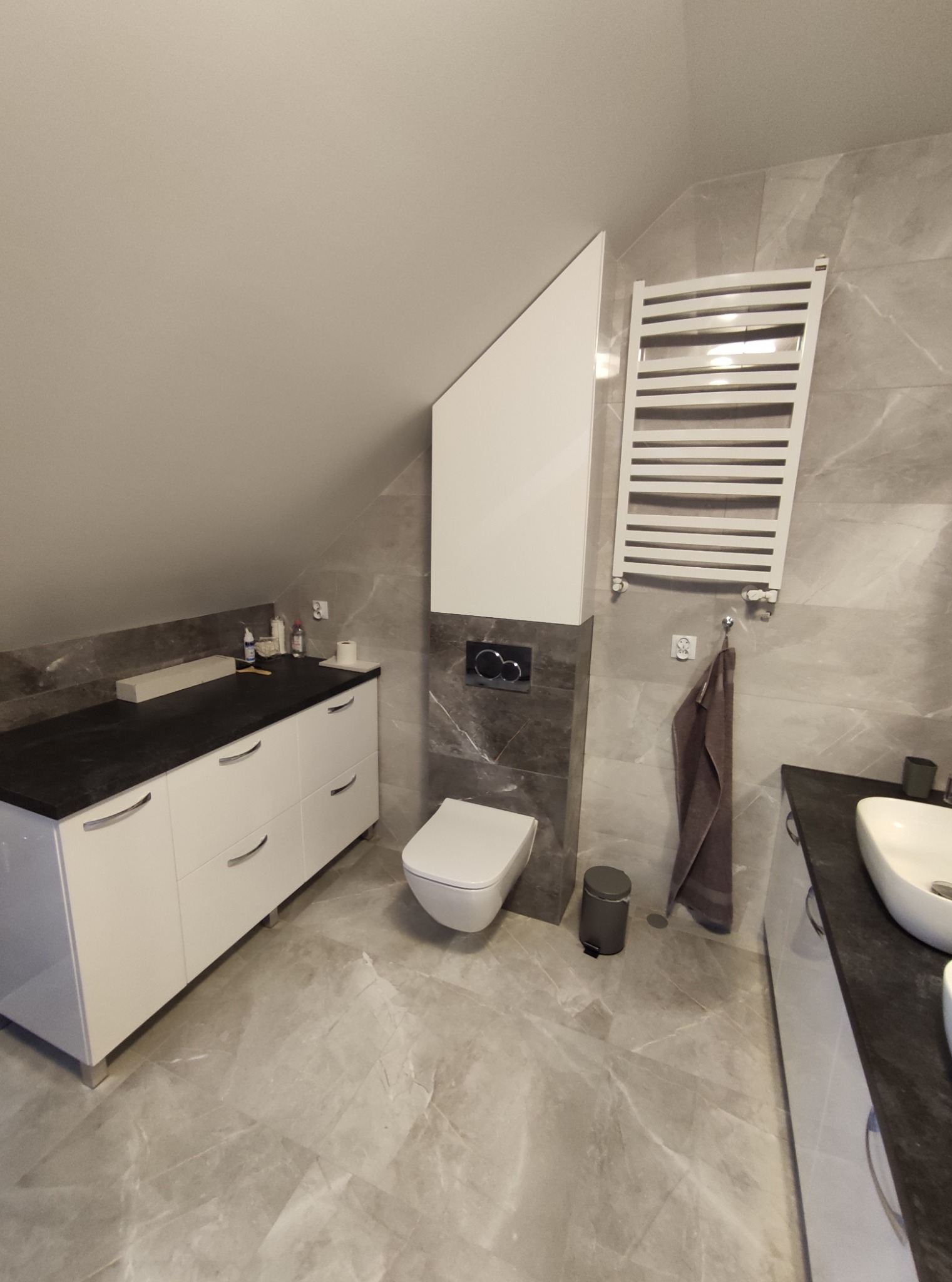 Nowoczesna łazienka na poddaszu z szarymi marmurowymi płytkami, białymi szafkami z czarnym blatem, podwieszaną toaletą i grzejnikiem drabinkowym.