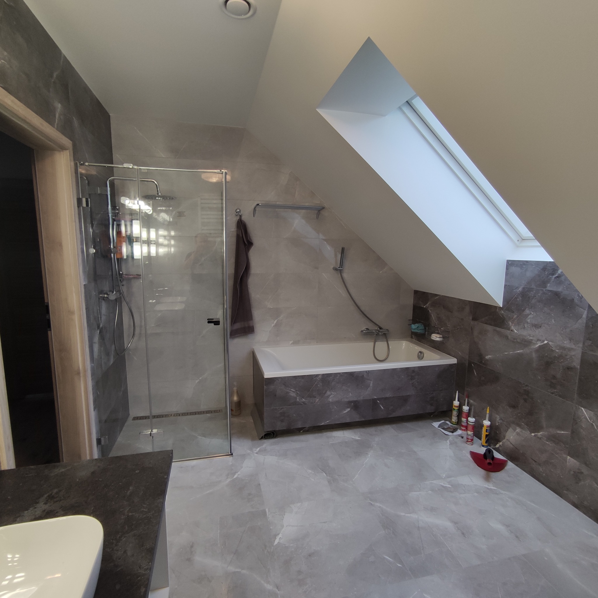Wnętrze łazienki na poddaszu z marmurową glazurą, kabiną prysznicową ze szklanymi drzwiami, wanną obudowaną marmurem, oknem dachowym i akcesoriami do silikonowania w rogu.