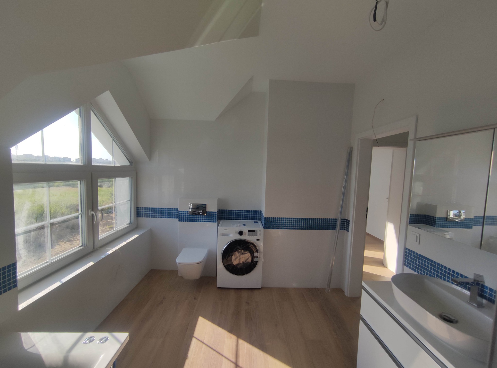 Jasna łazienka na poddaszu z oknem dachowym, białą armaturą, drewnopodobną podłogą i pasem niebieskich płytek dekoracyjnych, pralka w rogu.