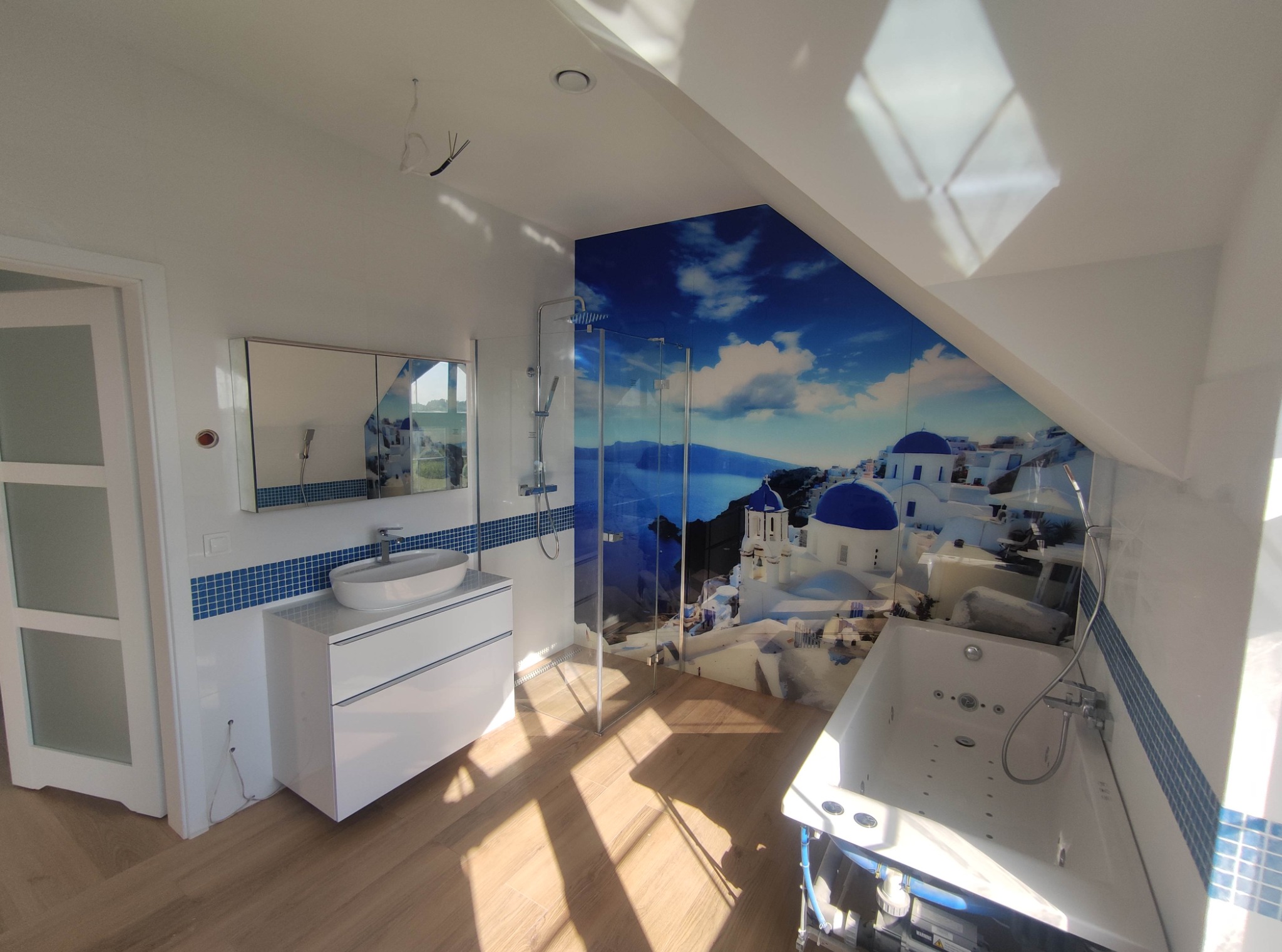 Jasna łazienka na poddaszu ze skośnym sufitem, białą zabudową umywalki, kabiną prysznicową i wanną z hydromasażem. Na ścianie fototapeta z motywem Santorini.