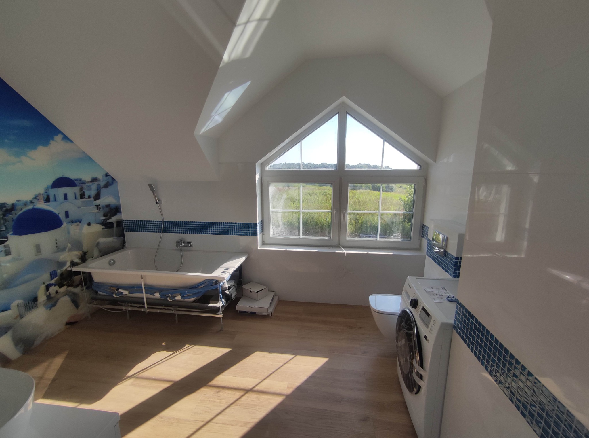 Jasna łazienka na poddaszu z oknem w kształcie trójkąta, białe płytki z niebieskim akcentem, drewnopodobna podłoga, wanna na metalowej konstrukcji, pralka i toaleta.