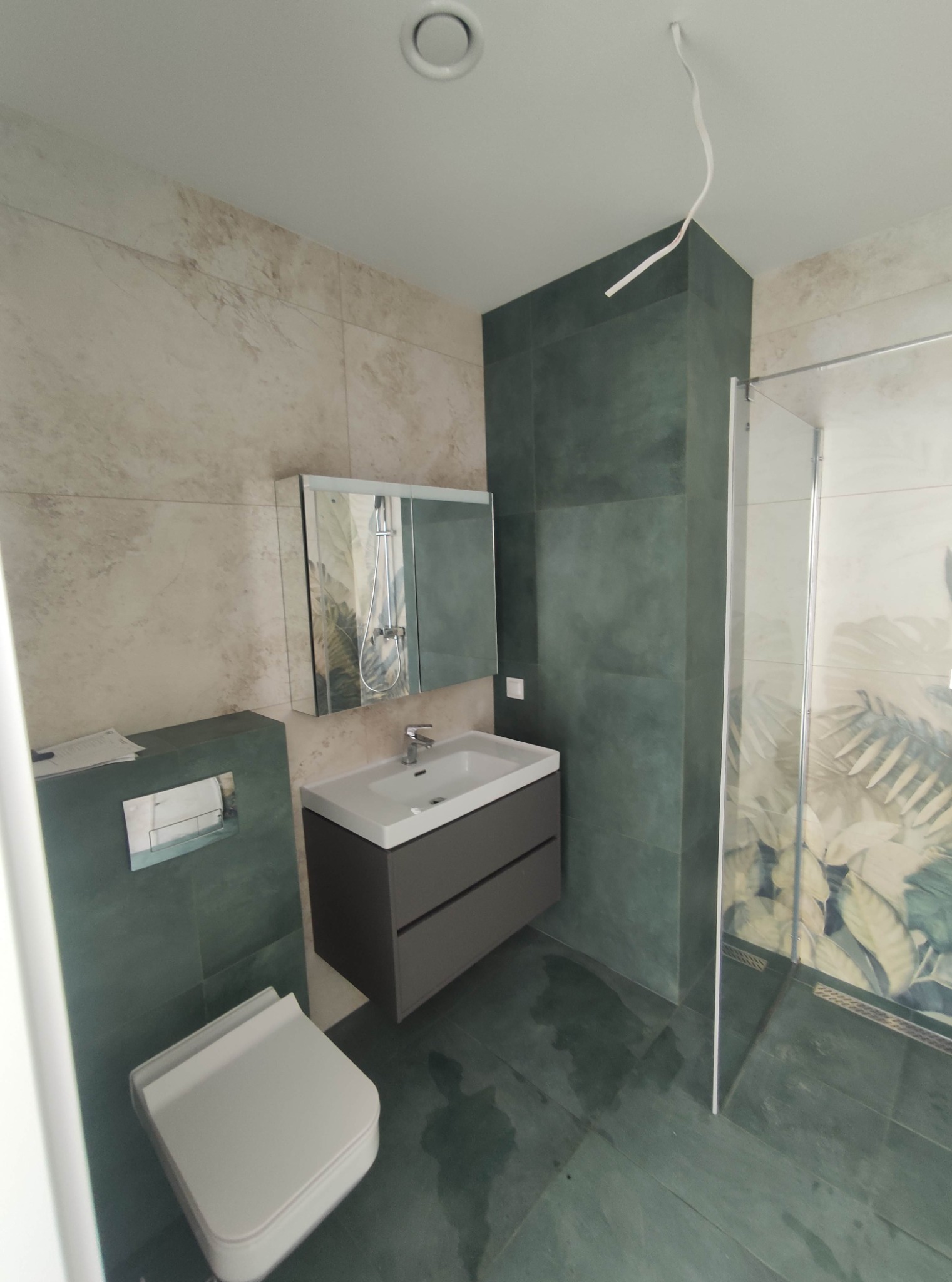 Wykończona łazienka z kabiną prysznicową z motywem roślinnym, umywalką z szafką w kolorze grafitowym, podwieszaną toaletą i zielonymi płytkami na podłodze i ścianach, z wystającym przewodem...