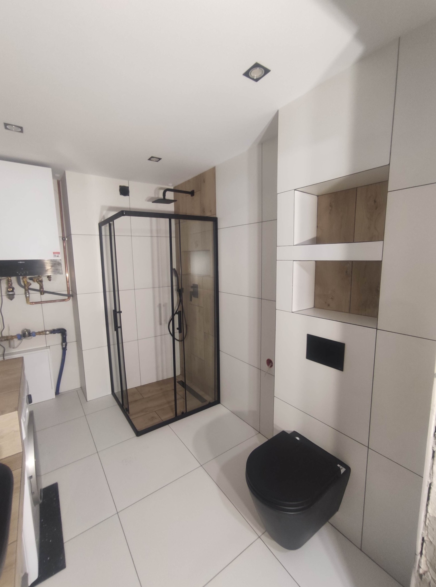 Nowoczesna łazienka z kabiną prysznicową w czarnej ramie, podwieszaną toaletą w czarnym kolorze, białymi płytkami i drewnianymi akcentami we wnękach ściennych.