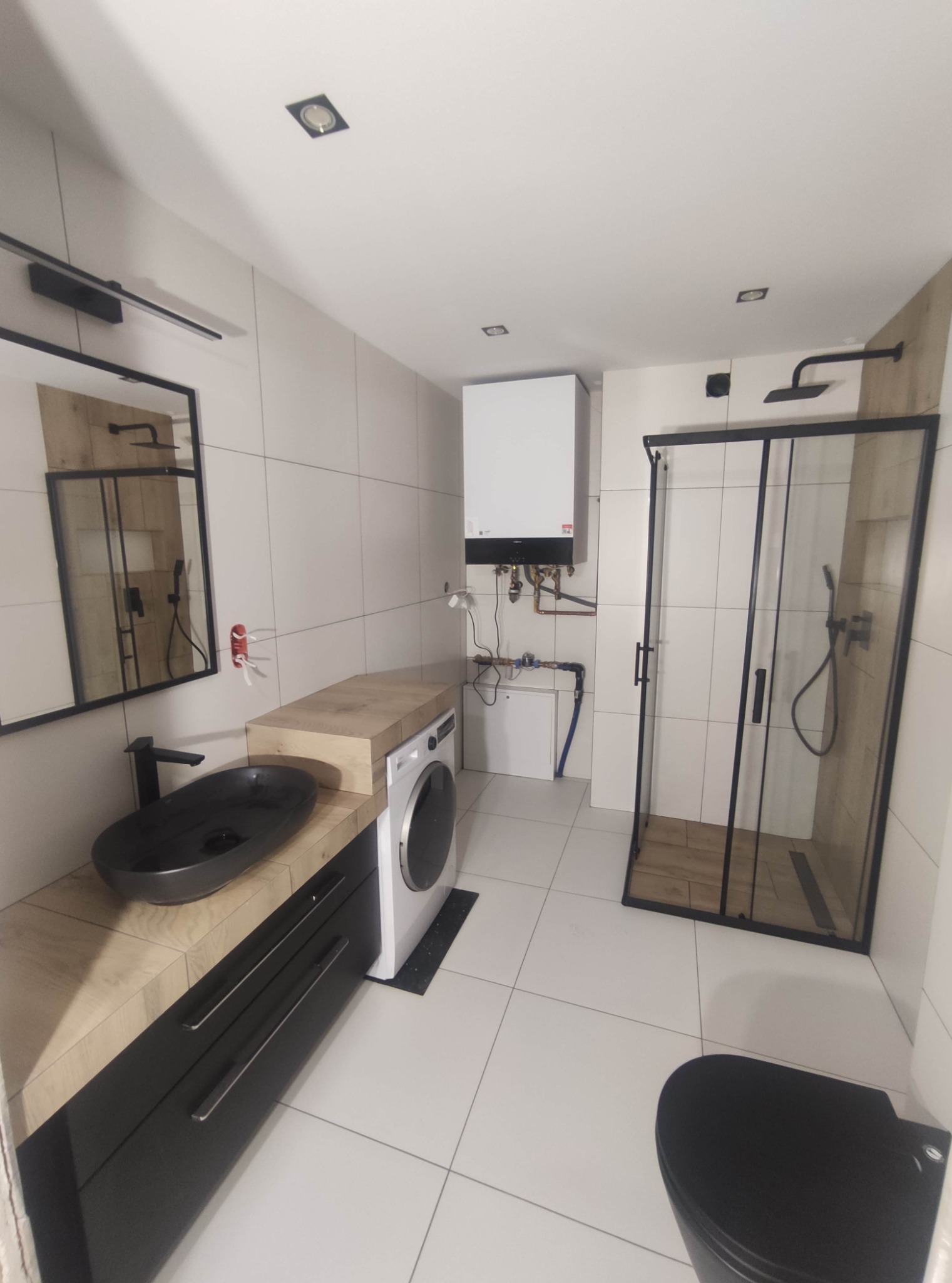 Nowoczesna łazienka w Żorach z kabiną prysznicową typu walk-in z czarnymi profilami, umywalką nablatową w kolorze czarnym, pralką oraz grzejnikiem, wykończona białymi płytkami.