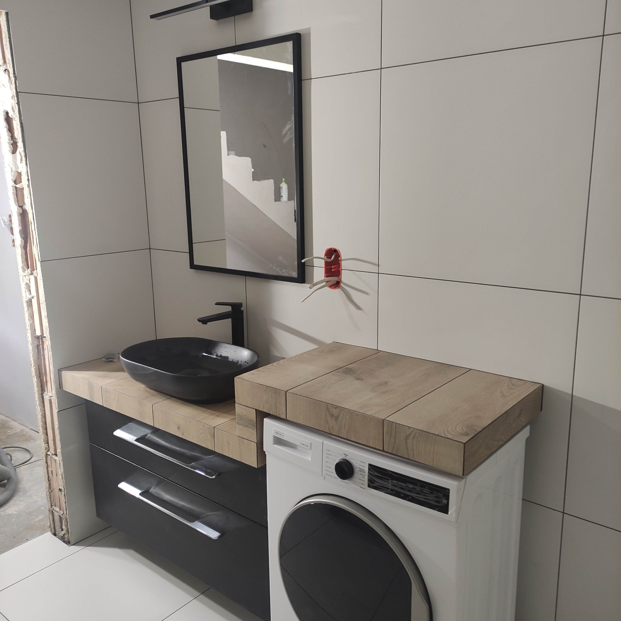Nowoczesna łazienka w trakcie wykańczania: czarna umywalka nablatowa, szafka z szufladami, pralka z drewnopodobnym blatem, białe płytki ścienne, widoczne kable elektryczne i niedokończone obrzeże.