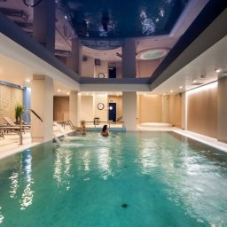 Sufity Napinane - Arkon System - Błękitny, lustrzany sufit w eleganckim basenie krytym z jacuzzi i dyszami wodnymi, oświetlony ciepłym światłem, z widocznymi osobami w wodzie.