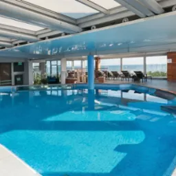 Błękitny basen wewnątrz budynku z widokiem na morze przez panoramiczne okna, oświetlony sufitem z przezroczystymi panelami, z drewnianymi elementami wykończenia i leżakami w tle.