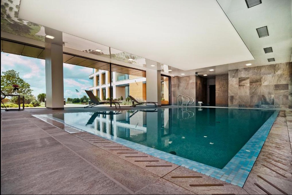 Kryty basen z turkusową wodą, szarymi płytkami i błyszczącym, białym sufitem z wbudowanym oświetleniem. Przez duże okno widoczny budynek i zieleń.