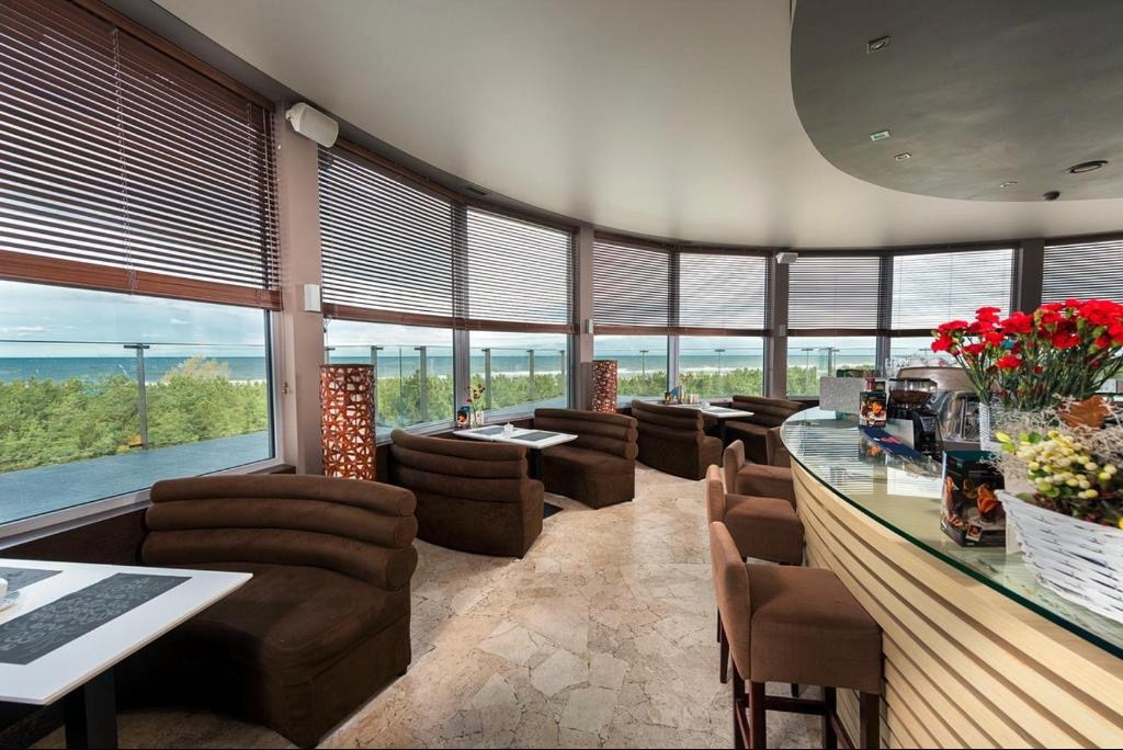 Wnętrze kawiarni z panoramicznym widokiem na morze, brązowe żaluzje poziome na oknach, brązowe sofy w kształcie kabin, marmurowa podłoga i szklany bar.