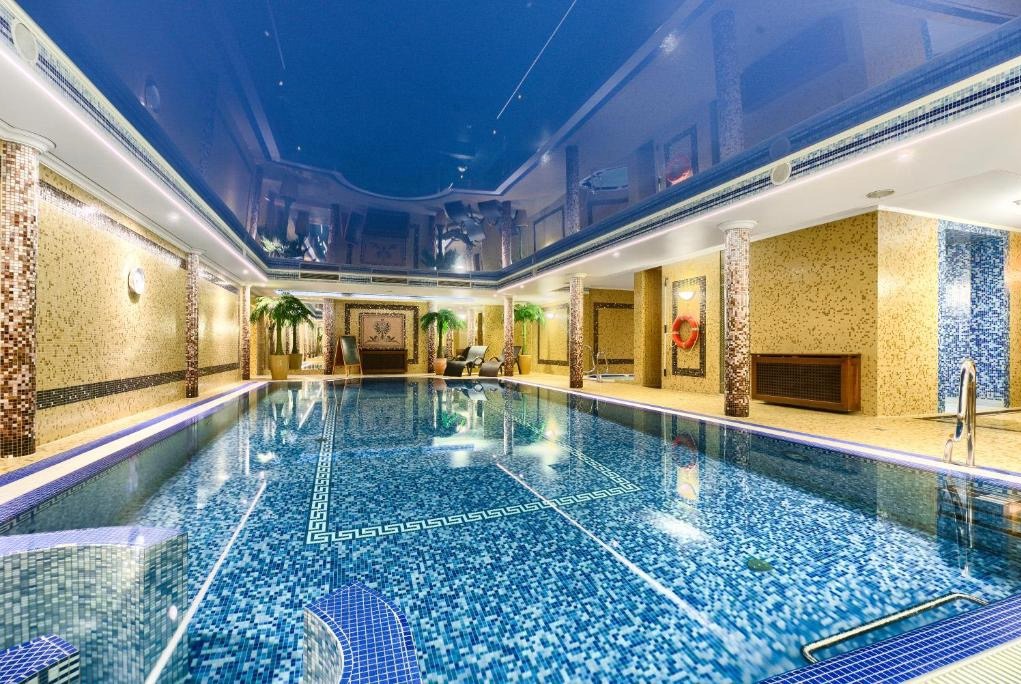Błękitny, błyszczący sufit napinany z refleksami w luksusowym basenie krytym, ściany wyłożone mozaiką, dekoracyjne kolumny i rośliny doniczkowe.