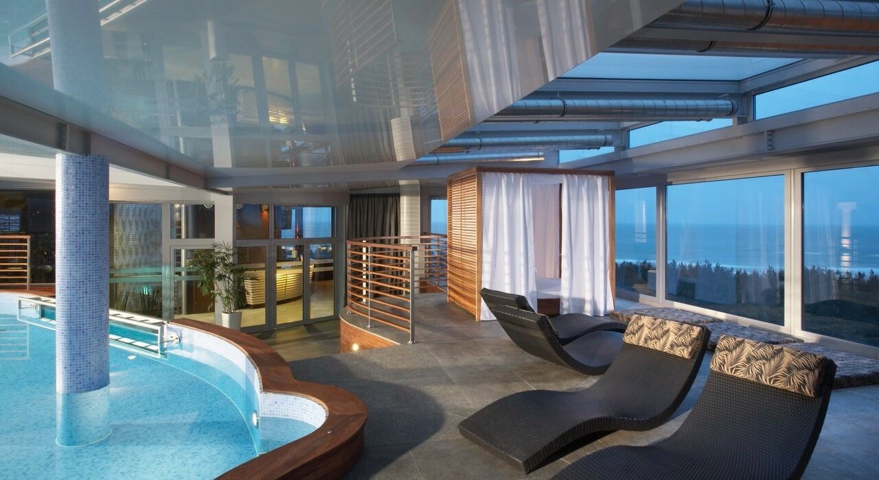 Luksusowe wnętrze spa z basenem, leżakami i widokiem na morze przez duże okna, akcentowane błyszczącym, refleksyjnym stropem.