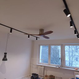 Vitek Vitek - Wnętrze pokoju w trakcie remontu z widocznymi narzędziami malarskimi, listwami drewnianymi na podłodze, lampą wiszącą i szynoprzewodami z reflektorami na suficie oraz wentylatorem sufitowym.