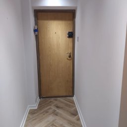 Vitek Vitek - Korytarz po remoncie z drewnopodobną podłogą ułożoną w jodełkę, świeżo pomalowane ściany na jasny kolor, nowe listwy przypodłogowe i drzwi wejściowe z ciemną futryną.