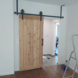 Vitek Vitek - Drewniane drzwi przesuwne w stylu rustykalnym z czarnym systemem mocowania na szynie, widoczne wnętrze mieszkania z podłogą i drabina malarska.