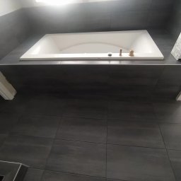 Vitek Vitek - Nowoczesna, wykafelkowana łazienka z wbudowaną białą wanną, widoczne elementy armatury przed montażem i instrukcja obsługi obok wanny, szare płytki na podłodze i ścianach, górne oświetlenie.