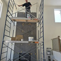 Vitek Vitek - Pracownik na rusztowaniu układa szare płytki kamienne na ścianie kominkowej w jasnym wnętrzu, widoczne krzyżyki dystansowe, kubełki z zaprawą, napis 'NU-WAY FLOORS' na rusztowaniu.